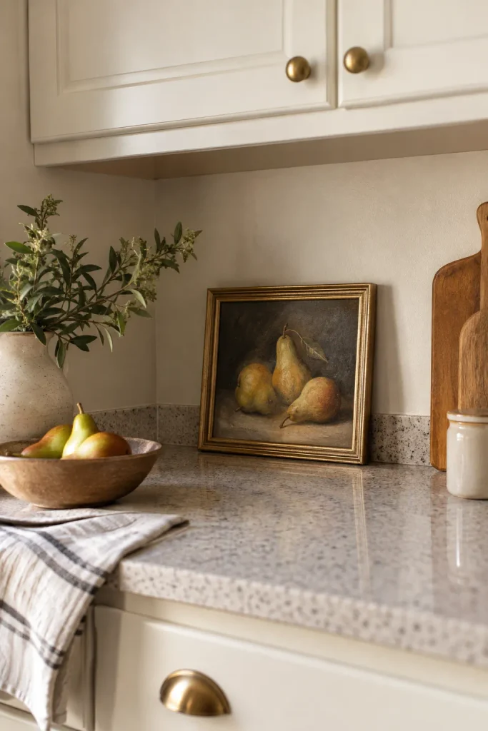 A framed pear still life leaning against the kitchen wall beside a wooden fruit bowl and cutting boards.
